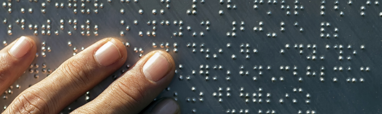 Foto em detalhe de dedos tocando uma placa de metal escrita em braile