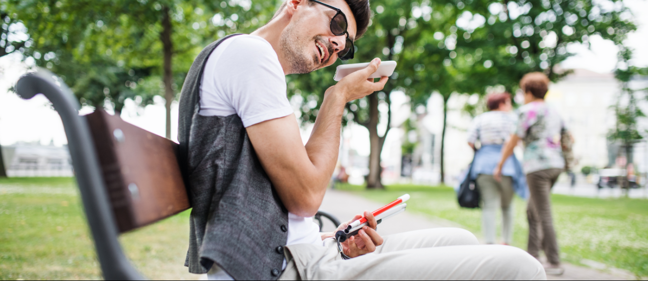 Homem sentado ao ar livre com o celular perto do rosto. Ele de óculos escuros, segura uma bengala e sorri.