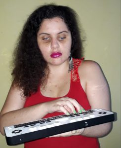 Sarah Barreto Marques, mulher branca, de cabelos castanhos, longos e cacheados, usa uma camisa regata vermelha. Com a mão direita, toca em uma linha Braille. O fundo da sala é amarelo claro.